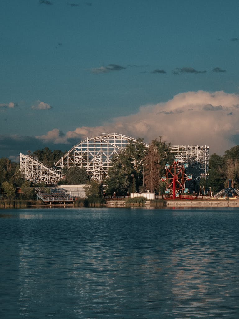 A picturesque view of a theme park with a roller coaster and ferris wheel by a body of water under a blue sky.